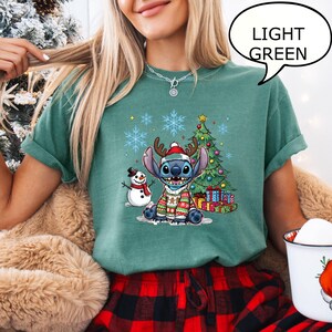 Puede incluir: Camiseta verde claro con un dise&ntilde;o de Stitch navide&ntilde;o. El dise&ntilde;o incluye a Stitch con un su&eacute;ter festivo y cuernos de reno, un mu&ntilde;eco de nieve, un &aacute;rbol de Navidad y regalos. El texto "LIGHT GREEN" est&aacute; en una burbuja.