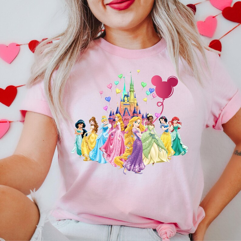 May include: Pink t-shirt with a graphic of Disney princesses and Cinderella's castle with Mickey Mouse ears balloon. The princesses are wearing their signature dresses and are standing in front of the castle.