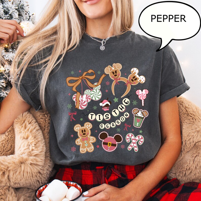 May include: Dark gray t-shirt with a festive design featuring gingerbread Mickey Mouse characters, candy canes, and the phrase "Tis the Season." The design includes bows, ornaments, and a coffee cup. The word "Pepper" is in a speech bubble.