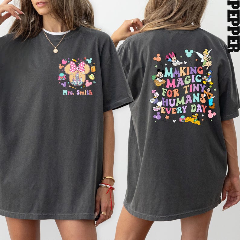 May include: Dark grey t-shirt with a Disney-themed design on the front and back. The front features a Minnie Mouse bow, castle, and "Mrs. Smith." The back says "Making Magic for Tiny Humans Every Day" with Disney characters.