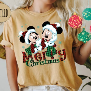May include: A mustard yellow t-shirt with a Christmas-themed design featuring Mickey Mouse and Minnie Mouse wearing red and black plaid scarves. The text "Merry Christmas" is printed below the characters.