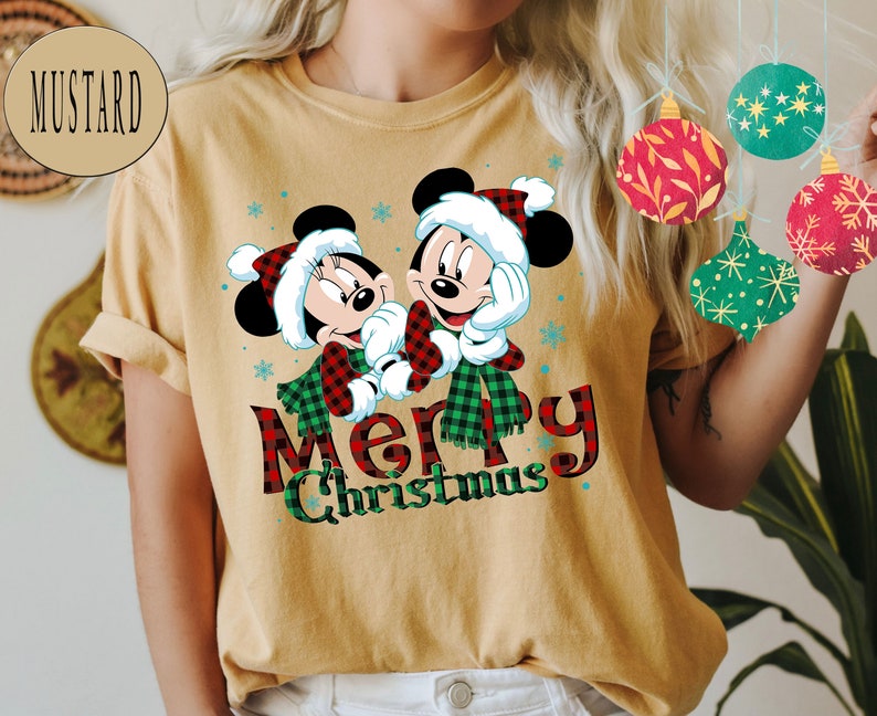 May include: A mustard yellow t-shirt with a Christmas-themed design featuring Mickey Mouse and Minnie Mouse wearing red and black plaid scarves. The text "Merry Christmas" is printed below the characters.
