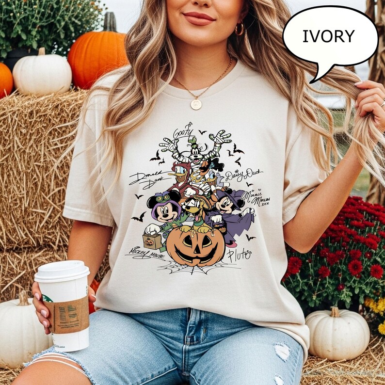 May include: Ivory-colored t-shirt featuring a Halloween-themed graphic with cartoon characters, including Mickey Mouse, Donald Duck, and Goofy. The design includes a carved pumpkin and bats. The shirt is worn by a person in blue jeans.