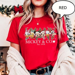 Puede incluir: Una camiseta roja con un dise&ntilde;o navide&ntilde;o que presenta personajes de Disney con atuendos festivos. La camiseta muestra el texto "MICKEY & CO." y "Est. 1928". Los personajes llevan cuernos de reno y bufandas. Un bocadillo dice "RED".