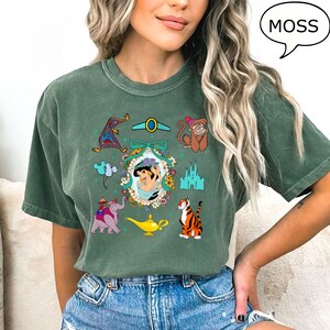 May include: A green t-shirt with a vintage-style print of various characters and objects from the Disney movie Aladdin. The print includes Jasmine, Abu, Rajah, the magic lamp, and the Cave of Wonders. The text "MOSS" is in a speech bubble in the upper right corner of the image.