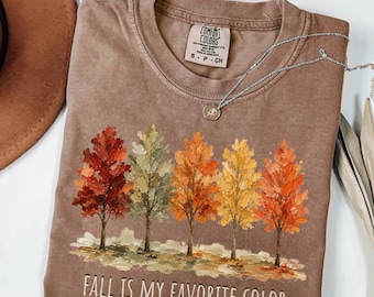 Puede incluir: Una camiseta de manga corta color espresso con un gráfico de cinco árboles con hojas de colores otoñales en rojo, verde, naranja y amarillo. El texto "FALL IS MY FAVORITE COLOR" está impreso en blanco debajo de los árboles. Un collar plateado descansa sobre la camiseta.