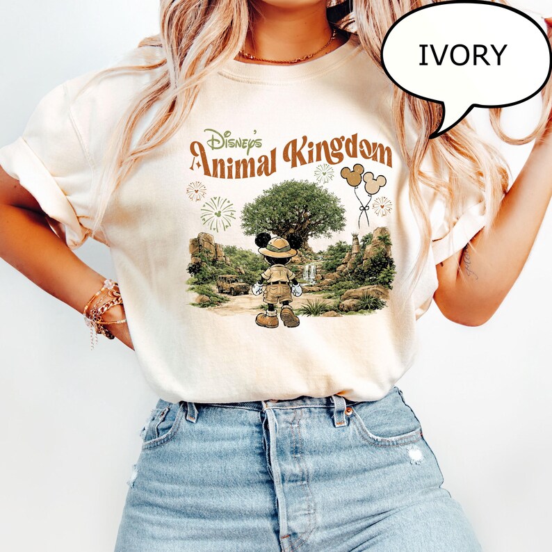 May include: Ivory-colored t-shirt featuring a graphic of Mickey Mouse in safari attire, set against a backdrop of Disney's Animal Kingdom. The design includes a large tree, waterfalls, and the park's logo. The shirt is a relaxed fit.