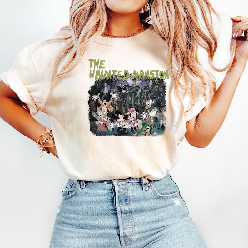 May include: A white t-shirt with a vintage-style graphic of Mickey Mouse and friends in front of the Haunted Mansion. The text "THE HAUNTED MANSION" is printed in green above the graphic.