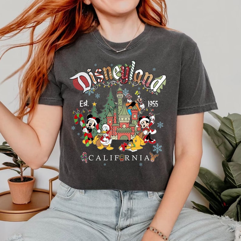 Puede incluir: Una camiseta negra con un logotipo de Disneyland de estilo vintage, con Mickey Mouse, Donald Duck, Goofy y Pluto frente al Castillo de la Bella Durmiente. El texto dice "Disneyland Est. 1955 California".