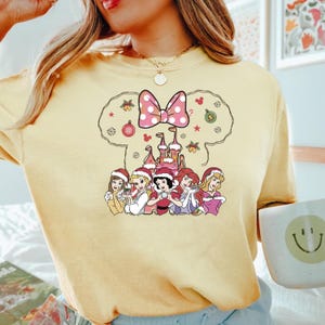 Puede incluir: Una camiseta amarilla con un dise&ntilde;o navide&ntilde;o que presenta un lazo rosa, un castillo y siete princesas Disney. Las princesas llevan gorros de Pap&aacute; Noel y sostienen art&iacute;culos navide&ntilde;os.
