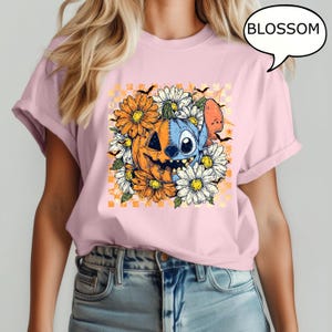 May include: Pink t-shirt featuring a Halloween-themed graphic. The design includes a smiling jack-o'-lantern, a blue alien character, and white and orange flowers. The word "BLOSSOM" is in a speech bubble.