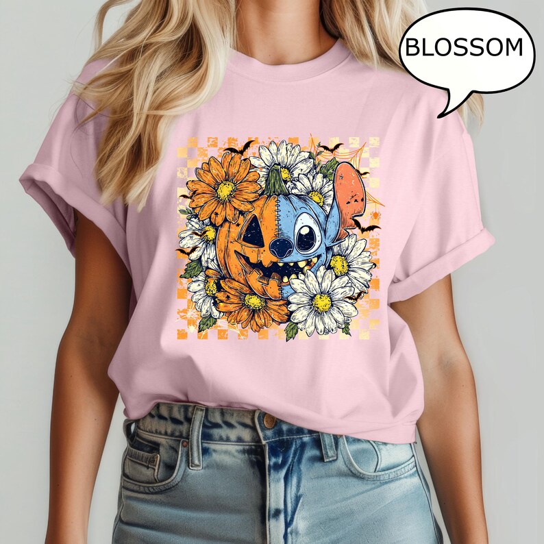 May include: Pink t-shirt featuring a Halloween-themed graphic. The design includes a smiling jack-o'-lantern, a blue alien character, and white and orange flowers. The word "BLOSSOM" is in a speech bubble.