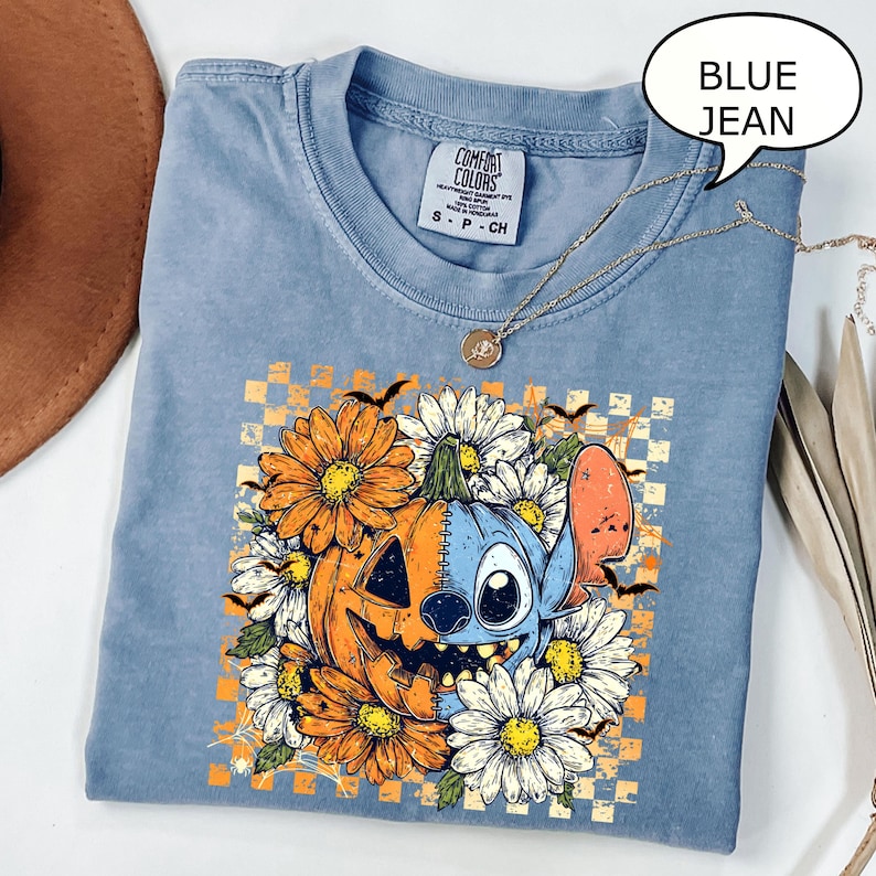 May include: A blue jean Comfort Colors t-shirt featuring a Halloween-themed graphic. The graphic includes a jack-o'-lantern, Stitch, daisies, and bats. A gold necklace is draped across the shirt.