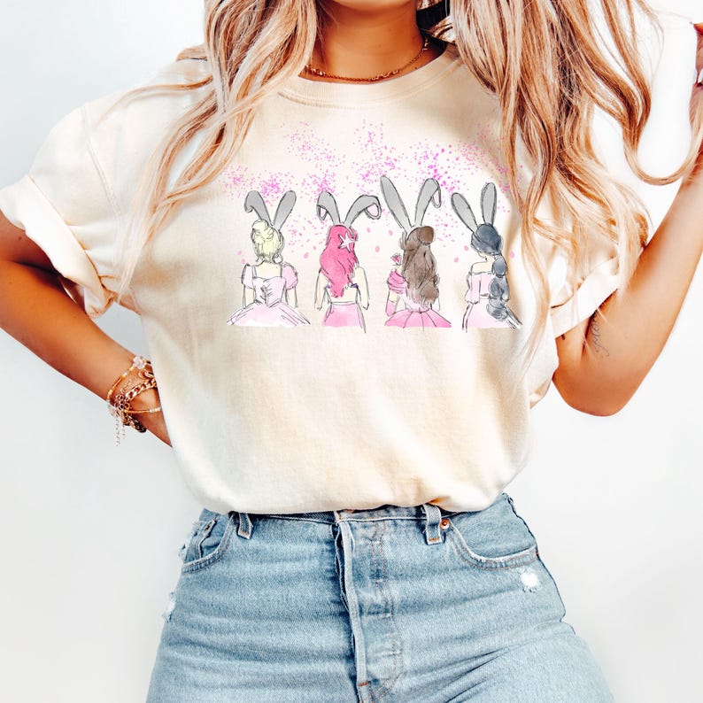 Puede incluir: Una camiseta blanca con un gr&aacute;fico de cuatro chicas con orejas de conejo y vestidos rosas. Las chicas est&aacute;n de espaldas al espectador y tienen el pelo de diferentes estilos.