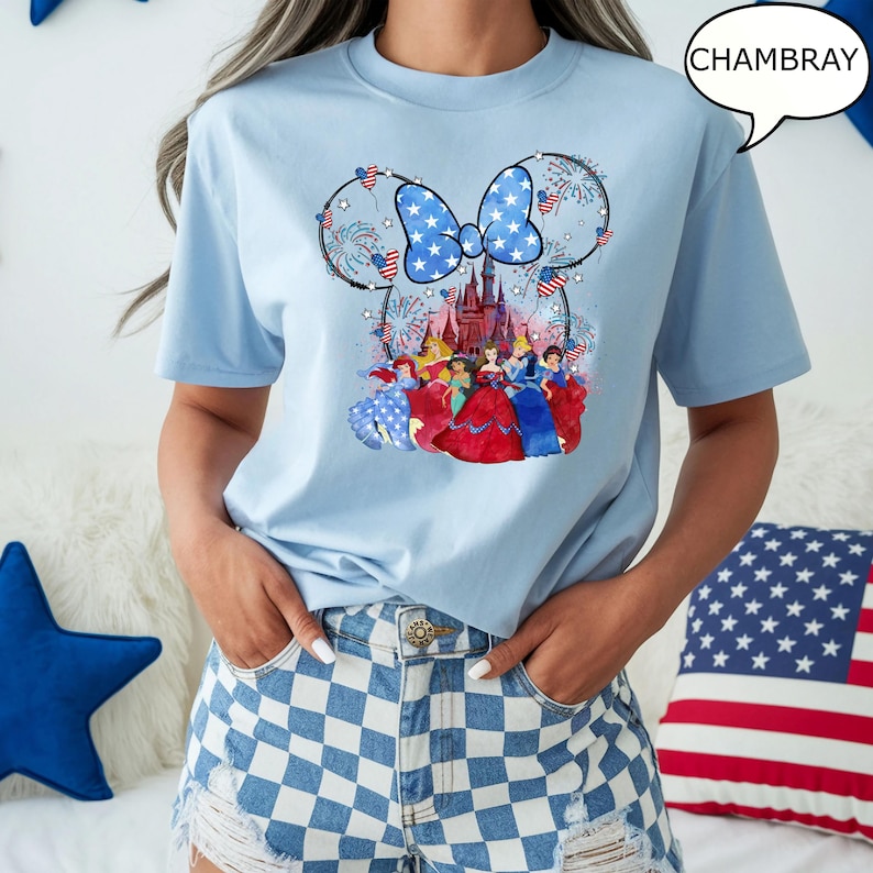 Puede incluir: Camiseta azul claro con un dise&ntilde;o patri&oacute;tico que presenta un lazo, fuegos artificiales y princesas de Disney. El lazo tiene un fondo azul con estrellas blancas. La camiseta tiene la palabra "CHAMBRAY" en una burbuja.