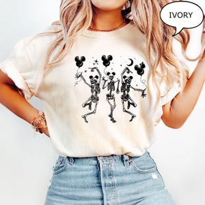 May include: Ivory-colored t-shirt featuring a black graphic of three dancing skeletons holding Mickey Mouse-shaped balloons. The design includes stars and a crescent moon. The shirt is a casual, Halloween-themed apparel item.