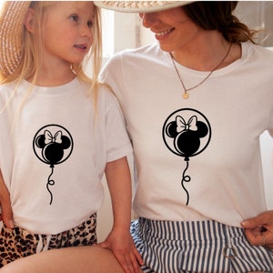 Pode incluir: Duas camisetas brancas com uma silhueta preta da cabe&ccedil;a da Minnie Mouse dentro de um bal&atilde;o. O bal&atilde;o tem um la&ccedil;o no topo e uma corda pendurada. As camisetas est&atilde;o sendo usadas por duas pessoas, uma adulta e uma crian&ccedil;a.