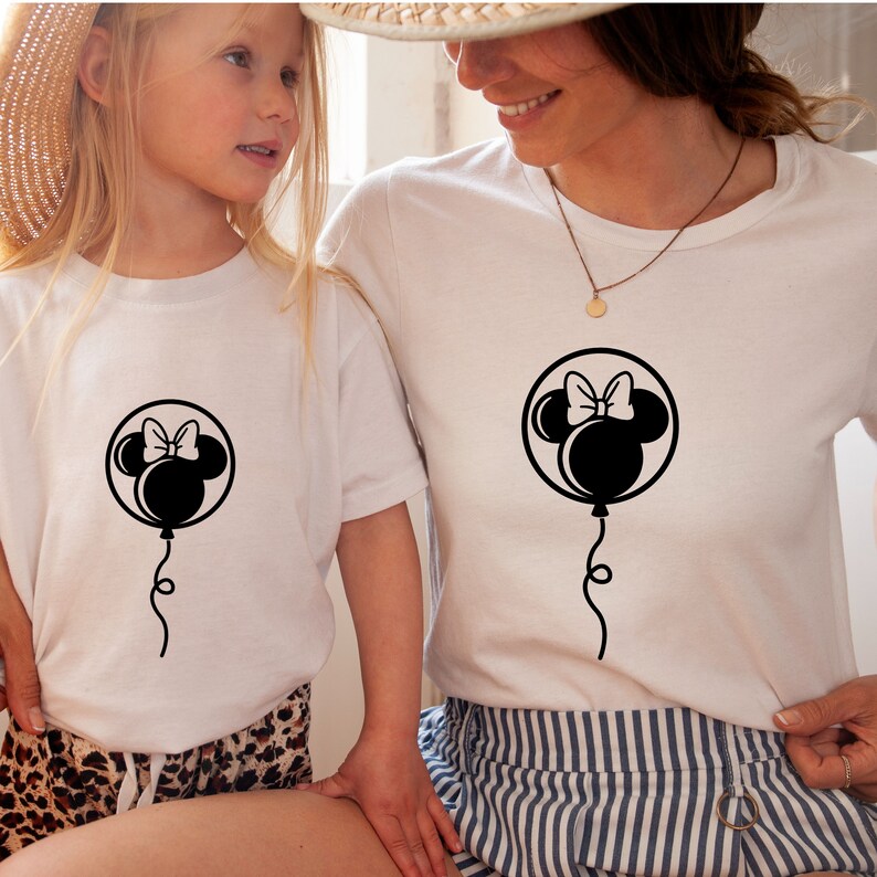 Pode incluir: Duas camisetas brancas com uma silhueta preta da cabe&ccedil;a da Minnie Mouse dentro de um bal&atilde;o. O bal&atilde;o tem um la&ccedil;o no topo e uma corda pendurada. As camisetas est&atilde;o sendo usadas por duas pessoas, uma adulta e uma crian&ccedil;a.