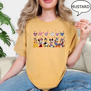 Puede incluir: Camiseta de color mostaza con un gr&aacute;fico de personajes de Disney sosteniendo globos en forma de coraz&oacute;n con patrones rojos, blancos y azules. Los personajes incluyen a Pluto, Mickey Mouse, Donald Duck, Minnie Mouse y Goofy. La palabra "MUSTARD" est&aacute; en una burbuja.