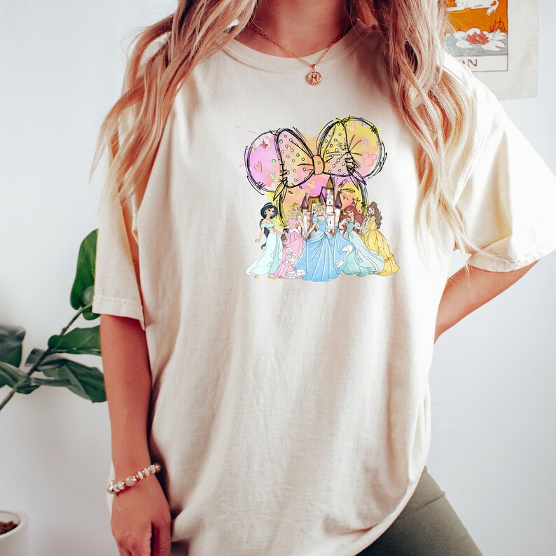 Puede incluir: Camiseta color crema con un dise&ntilde;o caprichoso de un gran lazo, un castillo y varias princesas. El lazo est&aacute; delineado en negro con acentos pastel rosa, amarillo y morado. Las princesas visten vestidos coloridos.