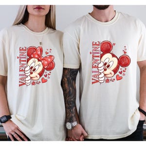 Puede incluir: Camisetas color crema con un dise&ntilde;o de San Valent&iacute;n. Una camiseta tiene un gr&aacute;fico de Minnie Mouse con un lazo rojo y la palabra "VALENTINE" en rojo. La otra camiseta tiene un gr&aacute;fico de Mickey Mouse con el mismo texto. Ambas camisetas tienen corazones rojos.
