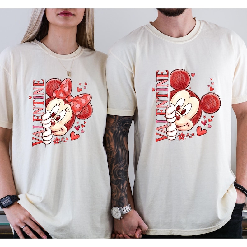 Puede incluir: Camisetas color crema con un dise&ntilde;o de San Valent&iacute;n. Una camiseta tiene un gr&aacute;fico de Minnie Mouse con un lazo rojo y la palabra "VALENTINE" en rojo. La otra camiseta tiene un gr&aacute;fico de Mickey Mouse con el mismo texto. Ambas camisetas tienen corazones rojos.