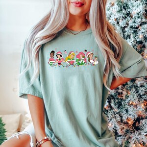 Puede incluir: Una camiseta azul claro con un gr&aacute;fico de seis princesas Disney vestidas con atuendos navide&ntilde;os y sosteniendo regalos. Las princesas est&aacute;n en fila con luces navide&ntilde;as y bastones de caramelo detr&aacute;s de ellas.