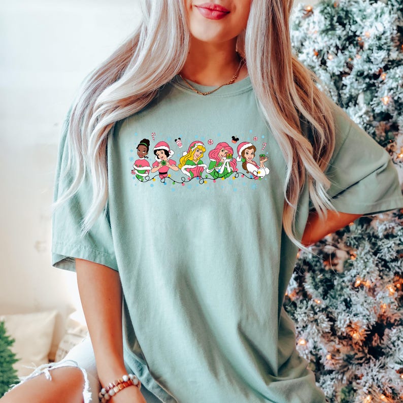 Puede incluir: Una camiseta azul claro con un gr&aacute;fico de seis princesas Disney vestidas con atuendos navide&ntilde;os y sosteniendo regalos. Las princesas est&aacute;n en fila con luces navide&ntilde;as y bastones de caramelo detr&aacute;s de ellas.