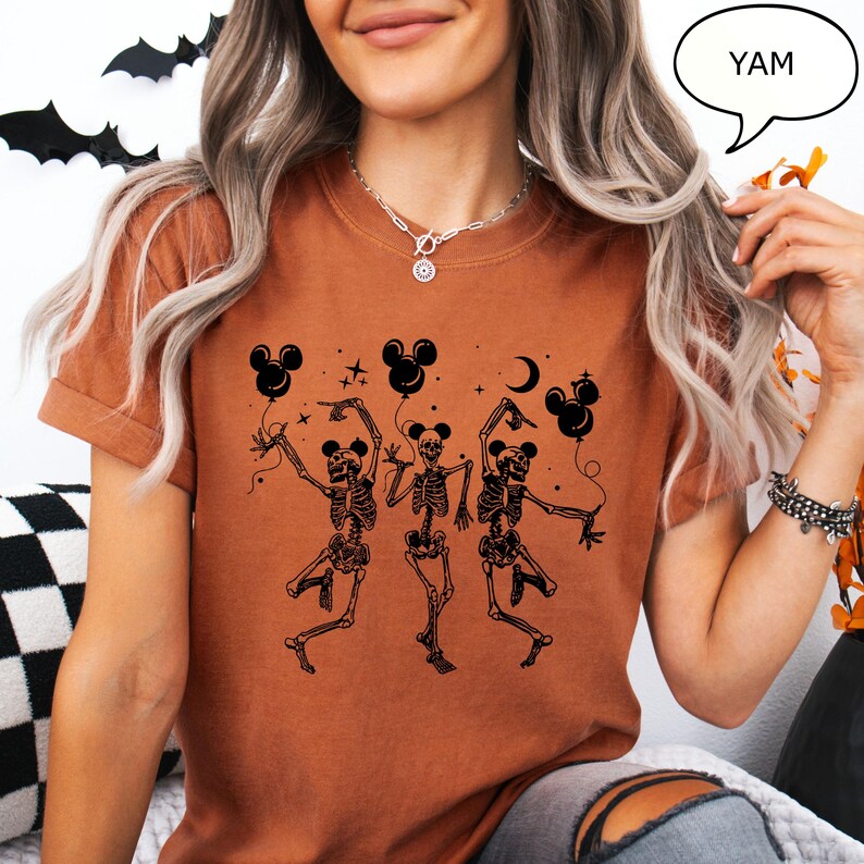 May include: A burnt orange t-shirt featuring a black graphic of three dancing skeletons holding Mickey Mouse-shaped balloons. The word "YAM" is in a speech bubble. The design includes stars and a crescent moon, perfect for Halloween.