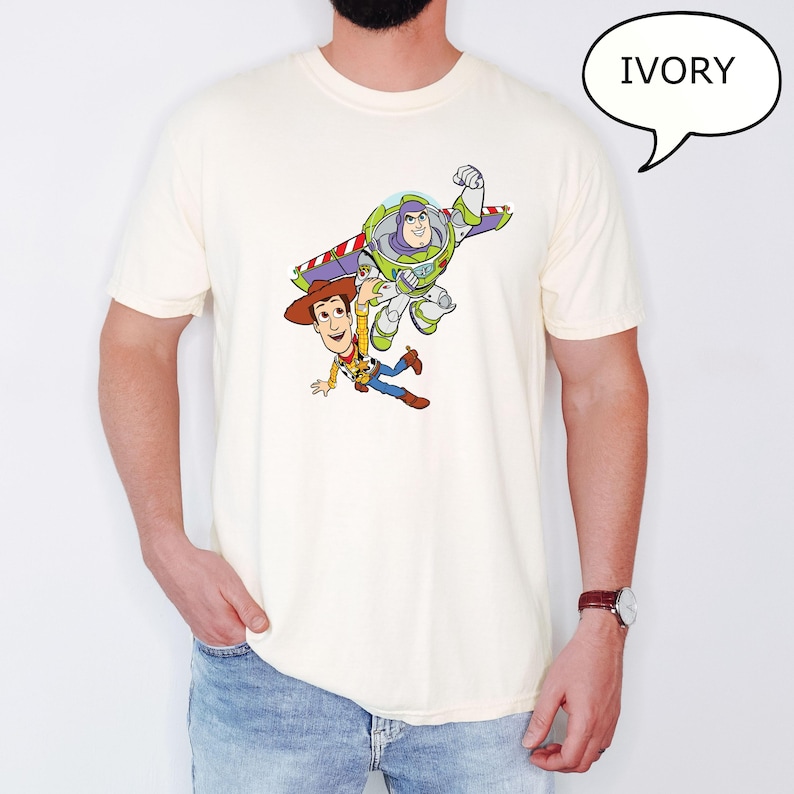 May include: Ivory-colored t-shirt featuring a graphic of Woody and Buzz Lightyear from Toy Story. Woody is wearing a cowboy hat and Buzz is in a spacesuit. The word "IVORY" is in a speech bubble.