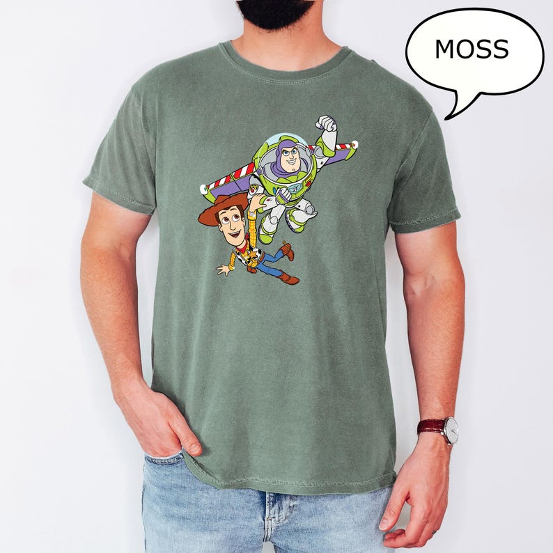 May include: A moss green t-shirt featuring a graphic of Buzz Lightyear and Woody from Toy Story. Buzz is in flight, and Woody is falling. A white speech bubble with the word "MOSS" is in the upper right corner.