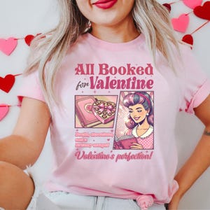 Puede incluir: Camiseta rosa con un dise&ntilde;o gr&aacute;fico que dice "All Booked for Valentine" con una imagen de dibujos animados de una mujer leyendo un libro y una caja de chocolates. El texto debajo de la imagen dice "Books, chocolates, and a dreamy escape! Valentine's perfection!"