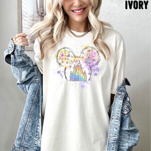 Puede incluir: Una camiseta blanca con una ilustraci&oacute;n colorida de una cabeza de Mickey Mouse con el ic&oacute;nico castillo de Disney en su interior. La ilustraci&oacute;n est&aacute; rodeada por un efecto de acuarela con salpicaduras de rosa, azul y amarillo.