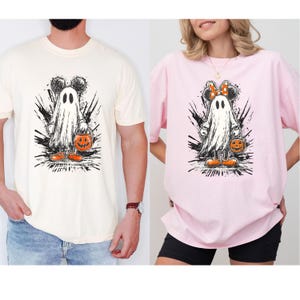 May include: Two t-shirts, one cream and one pink, featuring a Halloween-themed graphic. The graphic depicts a ghost with Mickey Mouse ears holding a jack-o'-lantern. The ghost is surrounded by black splatter designs, with orange shoes.