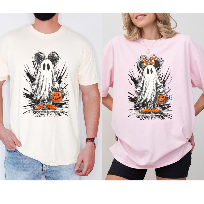 May include: Two t-shirts, one cream and one pink, featuring a Halloween-themed graphic. The graphic depicts a ghost with Mickey Mouse ears holding a jack-o'-lantern. The ghost is surrounded by black splatter designs, with orange shoes.