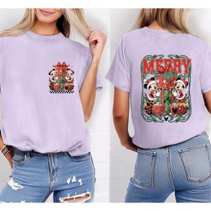 May include: A lilac t-shirt with a festive Christmas design featuring Mickey and Minnie Mouse. The front has a small graphic, while the back displays a larger "Merry Christmas" design with the characters, gifts, and decorative elements.