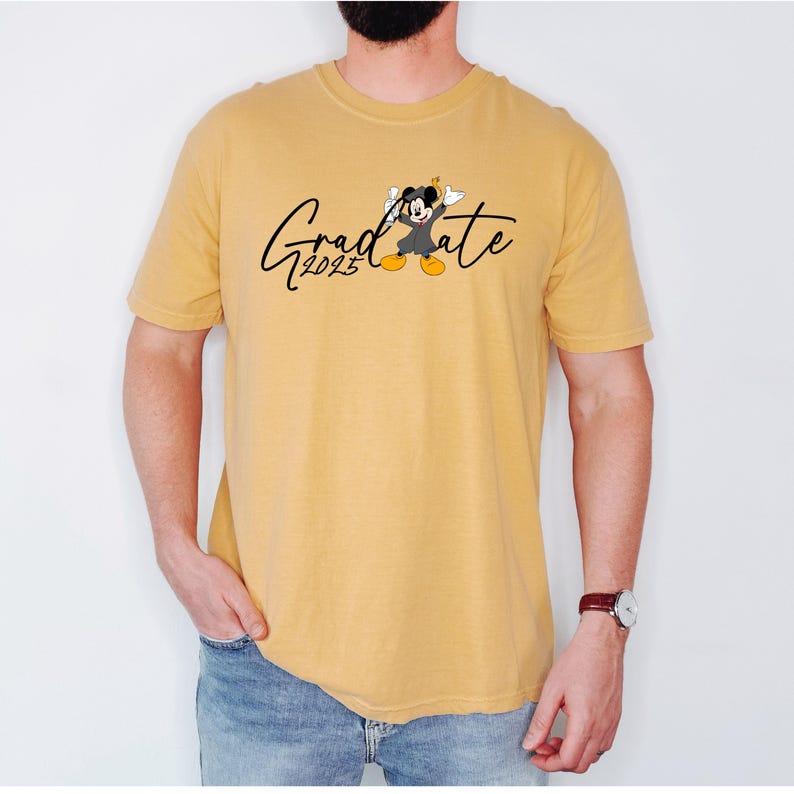 Puede incluir: Una camiseta amarilla con un gr&aacute;fico negro de Mickey Mouse con un gorro de graduaci&oacute;n y una bata. El texto "Grad 2025" est&aacute; impreso debajo del gr&aacute;fico.