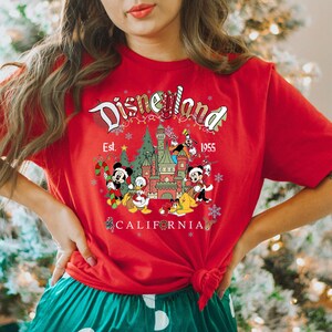 Puede incluir: Camiseta roja con un gr&aacute;fico de Disneyland con tem&aacute;tica navide&ntilde;a. El gr&aacute;fico presenta a Mickey Mouse, Donald Duck, Goofy, Pluto y el castillo de Disneyland. El texto "Disneyland Est. 1955 California" est&aacute; impreso en la camiseta.