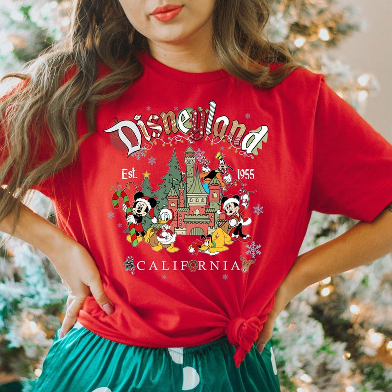 Puede incluir: Camiseta roja con un gr&aacute;fico de Disneyland con tem&aacute;tica navide&ntilde;a. El gr&aacute;fico presenta a Mickey Mouse, Donald Duck, Goofy, Pluto y el castillo de Disneyland. El texto "Disneyland Est. 1955 California" est&aacute; impreso en la camiseta.
