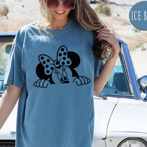 K&ouml;nnte beinhalten: Ein hellblaues T-Shirt mit einem schwarzen Aufdruck von Minnie Mouse, die &uuml;ber den Rand schaut. Der Aufdruck zeigt Minnies charakteristisches gepunktetes Schleifenband.