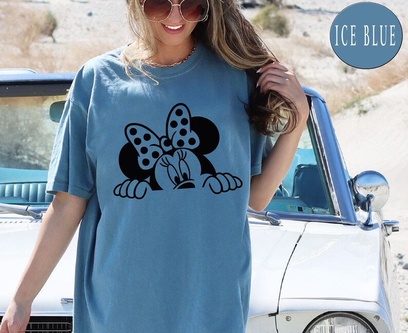 K&ouml;nnte beinhalten: Ein hellblaues T-Shirt mit einem schwarzen Aufdruck von Minnie Mouse, die &uuml;ber den Rand schaut. Der Aufdruck zeigt Minnies charakteristisches gepunktetes Schleifenband.