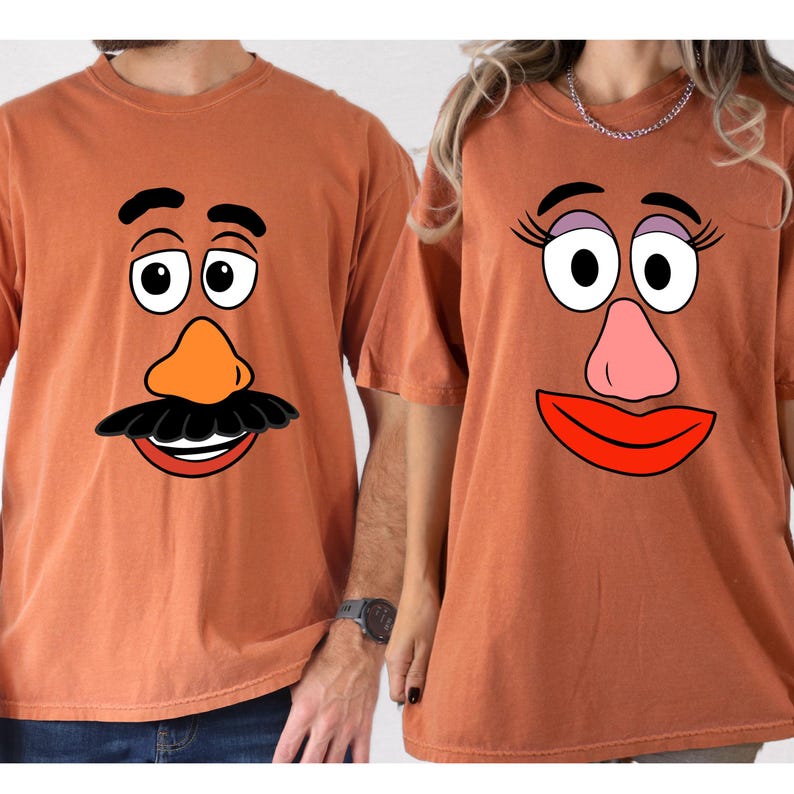 May include: Two orange-brown t-shirts with cartoon potato head faces. One shirt features a male potato head face with a black mustache and orange nose. The other shirt has a female potato head face with eyelashes and a pink nose.