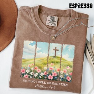 May include: A brown t-shirt with a faded look features a colourful illustration of a field of flowers with a cross on a hill in the background. The text "HE IS NOT HERE. HE HAS RISEN. Matthew 28:6" is printed on the shirt.