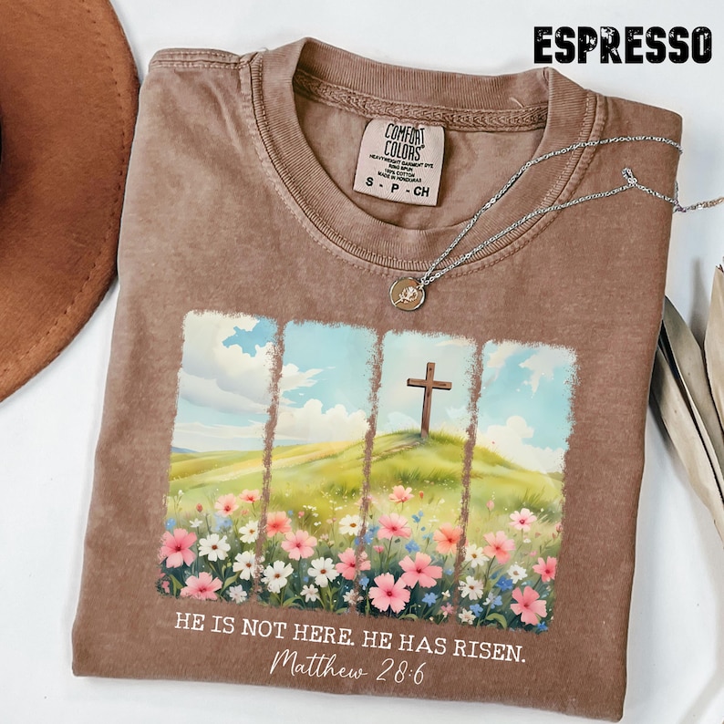 May include: A brown t-shirt with a faded look features a colourful illustration of a field of flowers with a cross on a hill in the background. The text "HE IS NOT HERE. HE HAS RISEN. Matthew 28:6" is printed on the shirt.