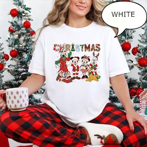 May include: White t-shirt with a colourful Christmas graphic featuring cartoon characters. The word "CHRISTMAS" is displayed across the top in a festive font. The characters include Mickey and Minnie Mouse, Goofy, and Pluto.