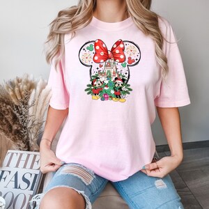 Puede incluir: Camiseta rosa con un lazo blanco y rojo de lunares, orejas de Mickey Mouse y una escena navide&ntilde;a con Mickey y Minnie Mouse de pie frente a un &aacute;rbol de Navidad y el castillo de Cenicienta.