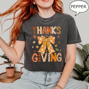 May include: Dark gray t-shirt with the words "THANKS GIVING" in a fall-themed design. The design includes a large orange bow, flowers, and leaves. The word "PEPPER" is in a speech bubble.
