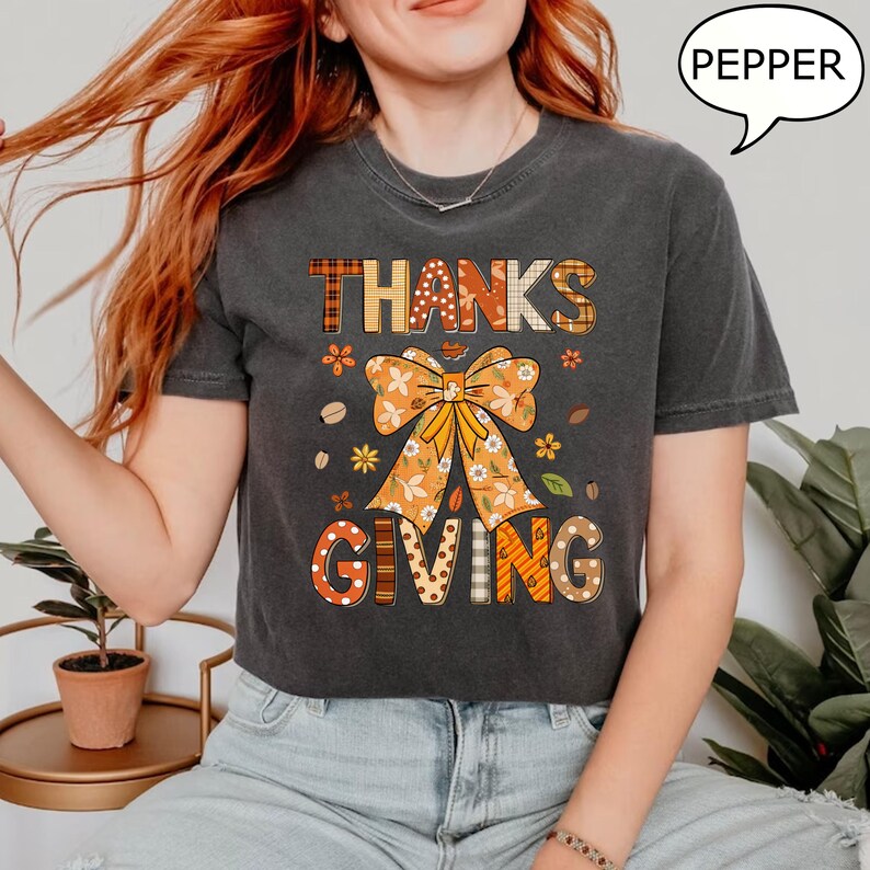 May include: Dark gray t-shirt with the words "THANKS GIVING" in a fall-themed design. The design includes a large orange bow, flowers, and leaves. The word "PEPPER" is in a speech bubble.