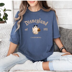 Puede incluir: Una camiseta azul con un logotipo vintage de Disneyland que presenta un fantasma con orejas de Mickey Mouse sosteniendo una calabaza. El texto dice "Disneyland est 1955 CALIFORNIA".