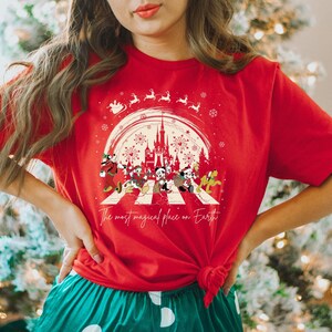 Puede incluir: Camiseta roja con un gr&aacute;fico blanco de Mickey Mouse y sus amigos caminando por un paso de cebra frente al Castillo de Cenicienta. El gr&aacute;fico tambi&eacute;n incluye copos de nieve, fuegos artificiales y el texto "The most magical place on Earth".